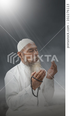 Muslim man with a beard wearing a white cap praying with prayer beads on his hands Muslim man with a beard wearing a white cap praying with prayer beads on his hands 102276113