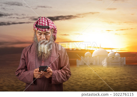 Muslim man with a beard wearing keffiyeh with agal using mobile phone 102276115