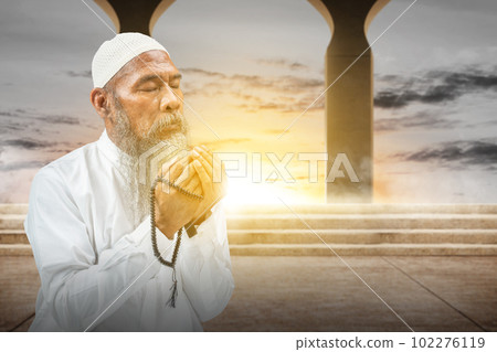 Muslim man with a beard wearing a white cap praying with prayer beads on his hands 102276119