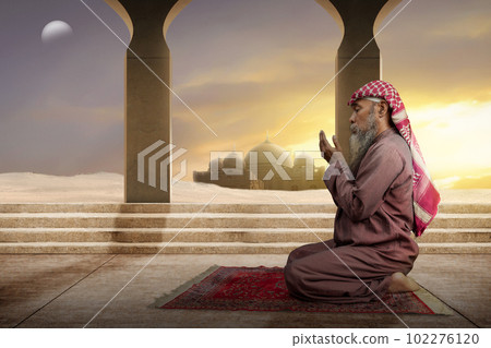 Muslim man with a beard wearing keffiyeh with agal in praying while raised hands 102276120