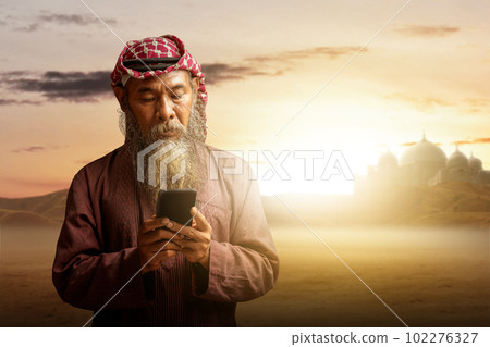 Muslim man with a beard wearing keffiyeh with agal using mobile phone 102276327