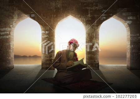 Muslim man with a beard wearing keffiyeh with agal sitting and reading the Quran on the prayer rug Muslim man with a beard wearing keffiyeh with agal sitting and reading the Quran on the prayer rug 102277203