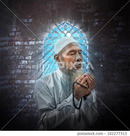 Muslim man with a beard wearing a white cap praying with prayer beads on his hands 102277313