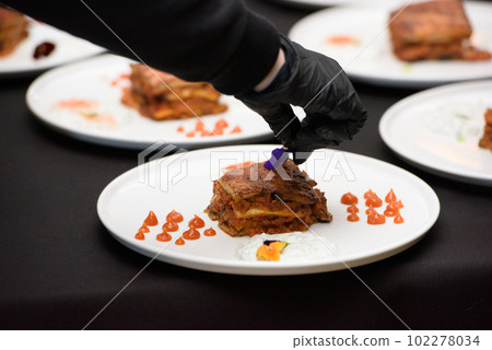 the chef in the restaurant preparing traditional greek moussaka in white plate on black tablecloth background the chef in the restaurant preparing traditional greek moussaka in white plate on black tablecloth background 102278034