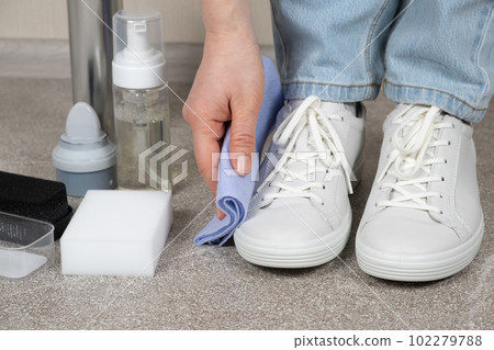 Cleaning white shoes, caring for sneakers. A woman wipes her leather shoes with a wet cloth from dust and dirt. Cleaning white shoes, caring for sneakers. A woman wipes her leather shoes with a wet cloth from dust and dirt. 102279788