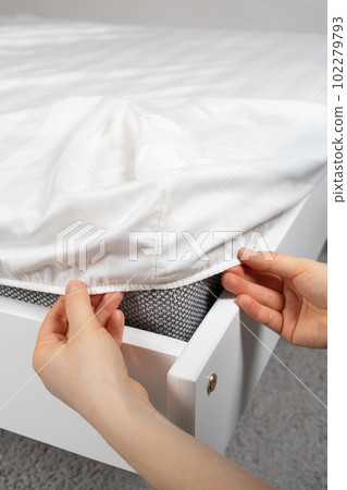 A white satin sheet with an elastic band is put on the mattress of the bed, a close-up of the hands. Bedclothes. A white satin sheet with an elastic band is put on the mattress of the bed, a close-up of the hands. Bedclothes. 102279793