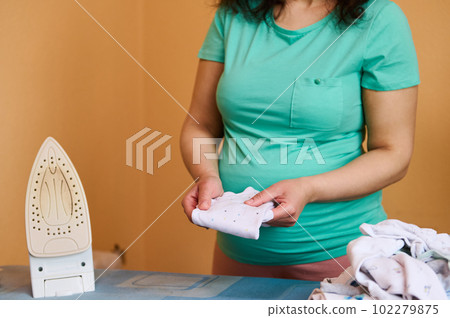 Close-up pregnant woman with big belly, expectant mother in turquoise t-shirt, stands by ironing board with steam iron and folding newborn clothes, preparing for childbirth. Happy moments of pregnancy 102279875