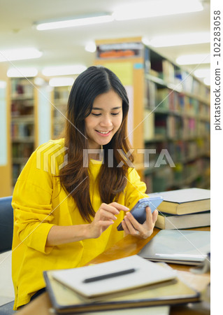Portrait of smiling young female student sitting at table in library with textbooks and using smartphone 102283058