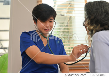 Male healthcare worker holding stethoscope checking heart and lungs of senior woman during home care visit Male healthcare worker holding stethoscope checking heart and lungs of senior woman during home care visit 102283089