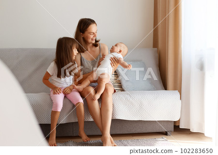 Portrait of happy family posing indoor in light living room while sitting on sofa near window with beige curtains, mother with two daughters at home, happy people. 102283650