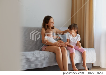 Happy family spending time together at home, mother holding little baby on arms and touching elder daughter hair, people spending time in light room while sitting on sofa. 102283651