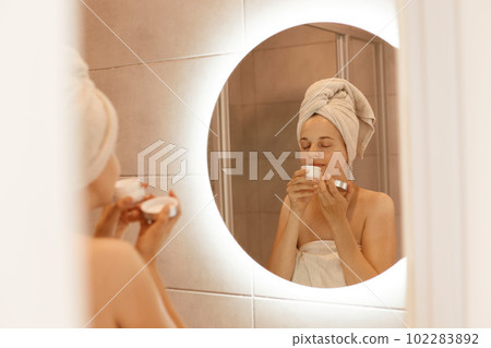 Young adult beautiful woman enjoying pleased cream odor, smelling cream before applying, standing with white towel on head in bathroom in front of mirror. 102283892