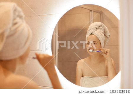 Attractive young adult female with white towel on her hair posing in front of the mirror in the bathroom and brushing her teeth, hygiene procedures after taking shower. 102283918