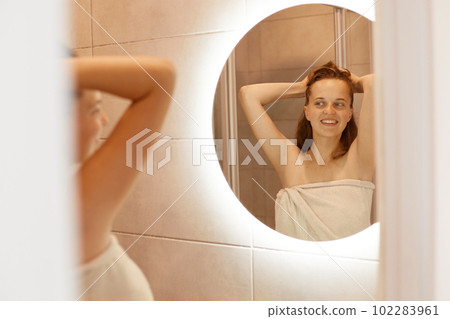 Smiling happy woman with perfect skin standing in front of mirror in bathroom with raised arms, morning routine, attractive female wrapped white towel. Smiling happy woman with perfect skin standing in front of mirror in bathroom with raised arms, morning routine, attractive female wrapped white towel. 102283961
