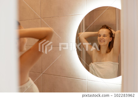 Indoor shot of smiling woman looking at her reflection in mirror with charming happy smile, standing with raised arms, female being wrapped in white towel. Indoor shot of smiling woman looking at her reflection in mirror with charming happy smile, standing with raised arms, female being wrapped in white towel. 102283962