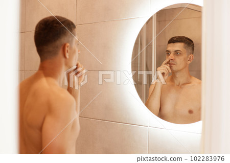 Indoor shot of young adult naked brunette male picking his nose in bathroom, standing with bare body, reflection in the mirror of man after taking shower. Indoor shot of young adult naked brunette male picking his nose in bathroom, standing with bare body, reflection in the mirror of man after taking shower. 102283976