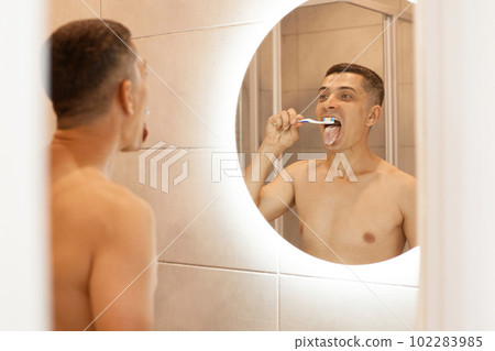 Portrait of brunette handsome man with bare torso standing in front of mirror in bathroom, brushing his tongue with toothbrush, keeps mouth widely opened, oral cavity hygiene. 102283985