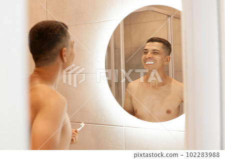 Extremely happy handsome man with bare torso standing in front of mirror in bathroom, brushing his teeth, looking at his teeth with toothpaste and smiling. 102283988