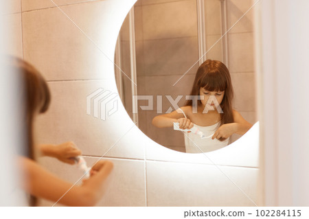 Portrait of cute dark haired child girl brushing teeth in bathroom, squeezing toothpaste out of a tube, standing in front of the mirror, wearing white sleeveless t shirt. 102284115