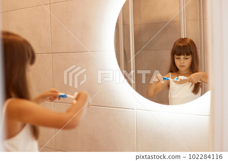 Indoor shot of charming little girl brushing teeth in bathroom, squeezing toothpaste out of a tube, standing in front of the mirror, having concentrated facial expression. Indoor shot of charming little girl brushing teeth in bathroom, squeezing toothpaste out of a tube, standing in front of the mirror, having concentrated facial expression. 102284116