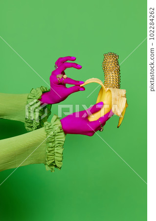 Portrait of colored female hands wearing rings holding gold banana over green studio background. Trash pop art, bright colors 102284262