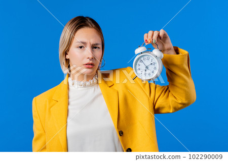 Confused woman holding alarm clock in hand. Blue background. Early 6 o'clock 102290009