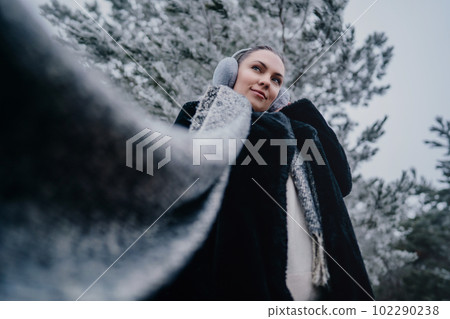 Trendy woman in fluffy earmuffs, long scarf on winter frozen forest background Trendy woman in fluffy earmuffs, long scarf on winter frozen forest background 102290238