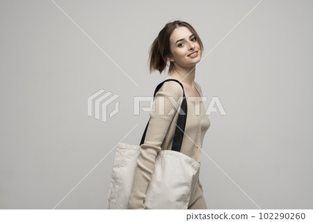 Brunette cheerful millennial woman holding white eco bag standing over white studio background. Lady holding flax shopper handbag. Fashion and ecology concept. Brunette cheerful millennial woman holding white eco bag standing over white studio background. Lady holding flax shopper handbag. Fashion and ecology concept. 102290260