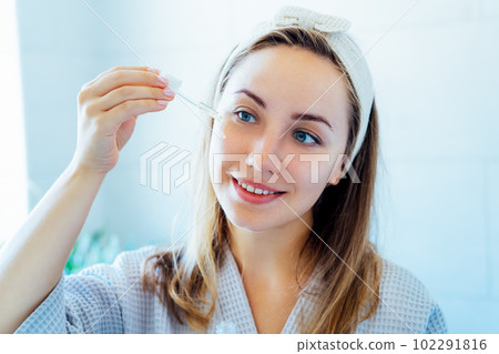 Young woman in bathrobe looking in the mirror, holding dropper with hyaluronic acid serum close to face and smiling. Skin hydrating. Cosmetic spa procedures. Beauty self-care at home. Selective focus. 102291816
