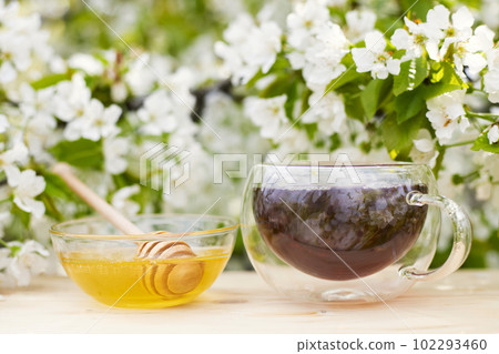 A glass cup of tea with honey in bowl on a wooden table on flowers background 102293460