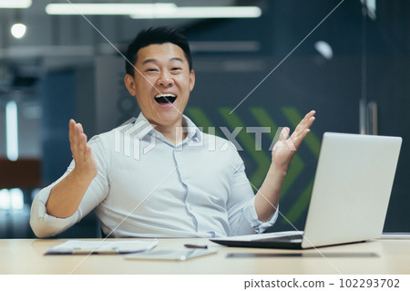 Happy Asian man sits in the office at the laptop and rejoices at the camera, spreads his hands, finished the project, made a successful deal, received a financial reward. 102293702