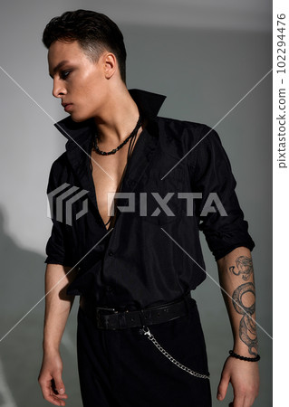 Profile portrait of a young attractive man in a black shirt and trousers, with a tattoo on his arm and fashion jewelry, short stylish hairstyle. Fashion sexy image. Gray background. Profile portrait of a young attractive man in a black shirt and trousers, with a tattoo on his arm and fashion jewelry, short stylish hairstyle. Fashion sexy image. Gray background. 102294476