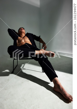 Wide angle portrait of a young brunette man in a black shirt and black trousers, with a muscular torso, sitting in a relaxed pose on a chair, looking up. Stylish studio shooting. Wide angle portrait of a young brunette man in a black shirt and black trousers, with a muscular torso, sitting in a relaxed pose on a chair, looking up. Stylish studio shooting. 102294477