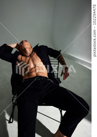 Wide angle portrait of a young brunette man in a black shirt and black trousers, with a muscular torso, sitting in a relaxed pose on a chair, looking up. Stylish studio shooting. Wide angle portrait of a young brunette man in a black shirt and black trousers, with a muscular torso, sitting in a relaxed pose on a chair, looking up. Stylish studio shooting. 102294478