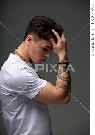Portrait of a young brunette boy in a white t-shirt with a snake tattoo on his arm, beads on his neck, a bracelet on his arm, fashionable short haircut. Contemporary urban fashion. Gray background. Portrait of a young brunette boy in a white t-shirt with a snake tattoo on his arm, beads on his neck, a bracelet on his arm, fashionable short haircut. Contemporary urban fashion. Gray background. 102294496