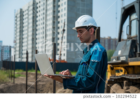 A male builder in a helmet stands against the background of a construction site with a laptop in his hands. A male builder in a helmet stands against the background of a construction site with a laptop in his hands. 102294522
