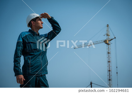 A builder in work clothes and a helmet stands on a construction site against the background of a construction crane.  102294681