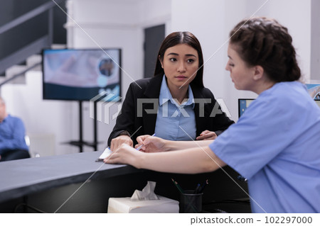 Nurse showing medical report to receptionist discussing expertise during checkup visit examination in hospital waiting room. Worker helping assistant with consultations and follow-up appointments. 102297000