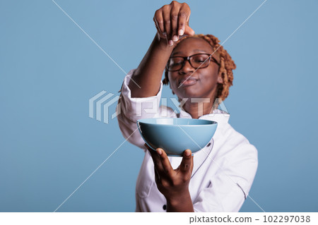 African American chef holding bowl of food while adding salt with style in front of the camera. Professional cook proud of new preparation in studio shot against blue background. African American chef holding bowl of food while adding salt with style in front of the camera. Professional cook proud of new preparation in studio shot against blue background. 102297038
