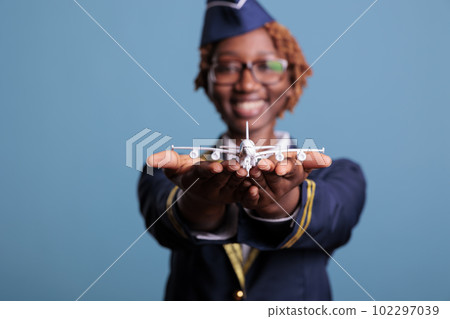 Funny flight attendant in uniform landing model airplane in hands, playing with small custom toy. Airplane crew member with passenger toy, studio shot against blue background. 102297039