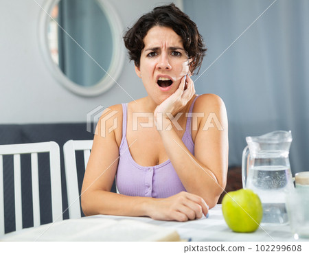 Young woman having toothache and touching cheek, sitting at table 102299608