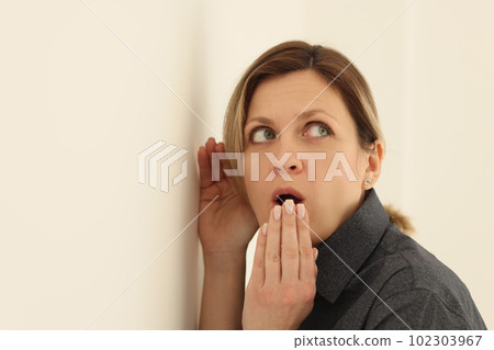 Curious woman puts ear close to beige wall covering mouth Curious woman puts ear close to beige wall covering mouth 102303967