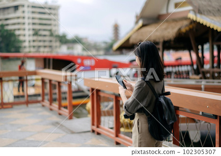 Young Asian woman backpack traveler using mobile phone in express boat pier on Chao Phraya River in Bangkok. 102305307