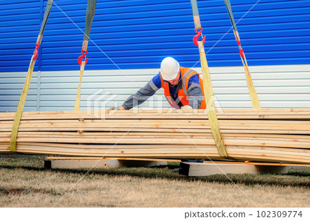 Slinger in helmet and signal vest unloads wooden boards on summer day outside on street Slinger in helmet and signal vest unloads wooden boards on summer day outside on street 102309774