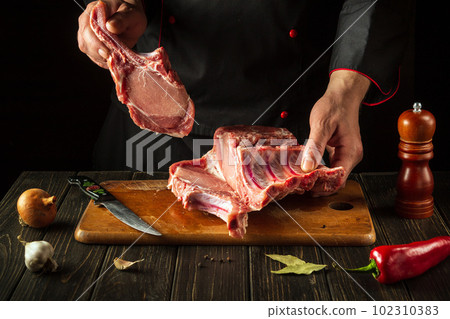 The chef is holding a piece of raw meat with a bone in the kitchen. Slicing pork ribs by the hands of a butcher. European cuisine The chef is holding a piece of raw meat with a bone in the kitchen. Slicing pork ribs by the hands of a butcher. European cuisine 102310383