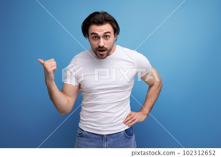 surprised brunette young man in a white t-shirt shows his index finger to the side on a studio 102312652