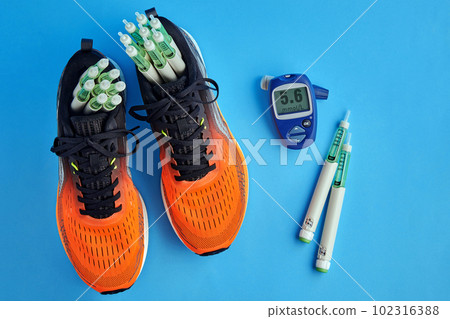 Orange running shoes with insulin syringe pens and a glucometer for measuring blood sugar surrounded by decorative red hearts on a blue background 102316388