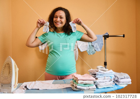 Happy ethnic pregnant woman holding newborn scrapers and steam iron, smiling cute, looking at camera, standing at ironing board with clean ironed baby clothes arranged in a stack. Pregnancy. Maternity Happy ethnic pregnant woman holding newborn scrapers and steam iron, smiling cute, looking at camera, standing at ironing board with clean ironed baby clothes arranged in a stack. Pregnancy. Maternity 102317728