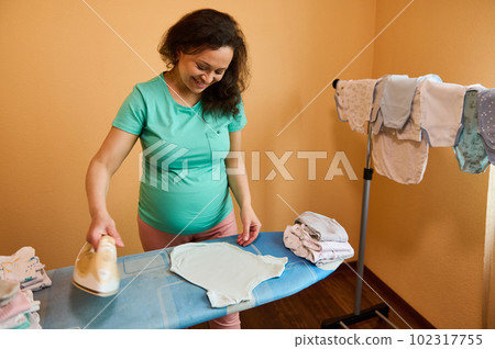 Smiling pregnant woman in home clothes, enjoying happy moments of her pregnancy and future maternity, ironing newborn bodysuit with steam iron. Household chore. Motherhood. People. Children. Lifestyle 102317755