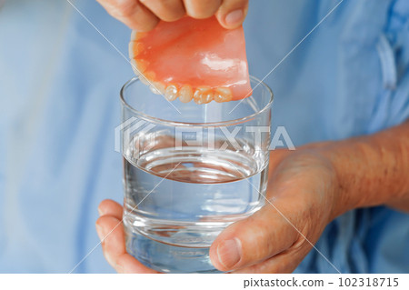 Asian senior woman patient clean teeth denture in a glass with solution for chew food. 102318715